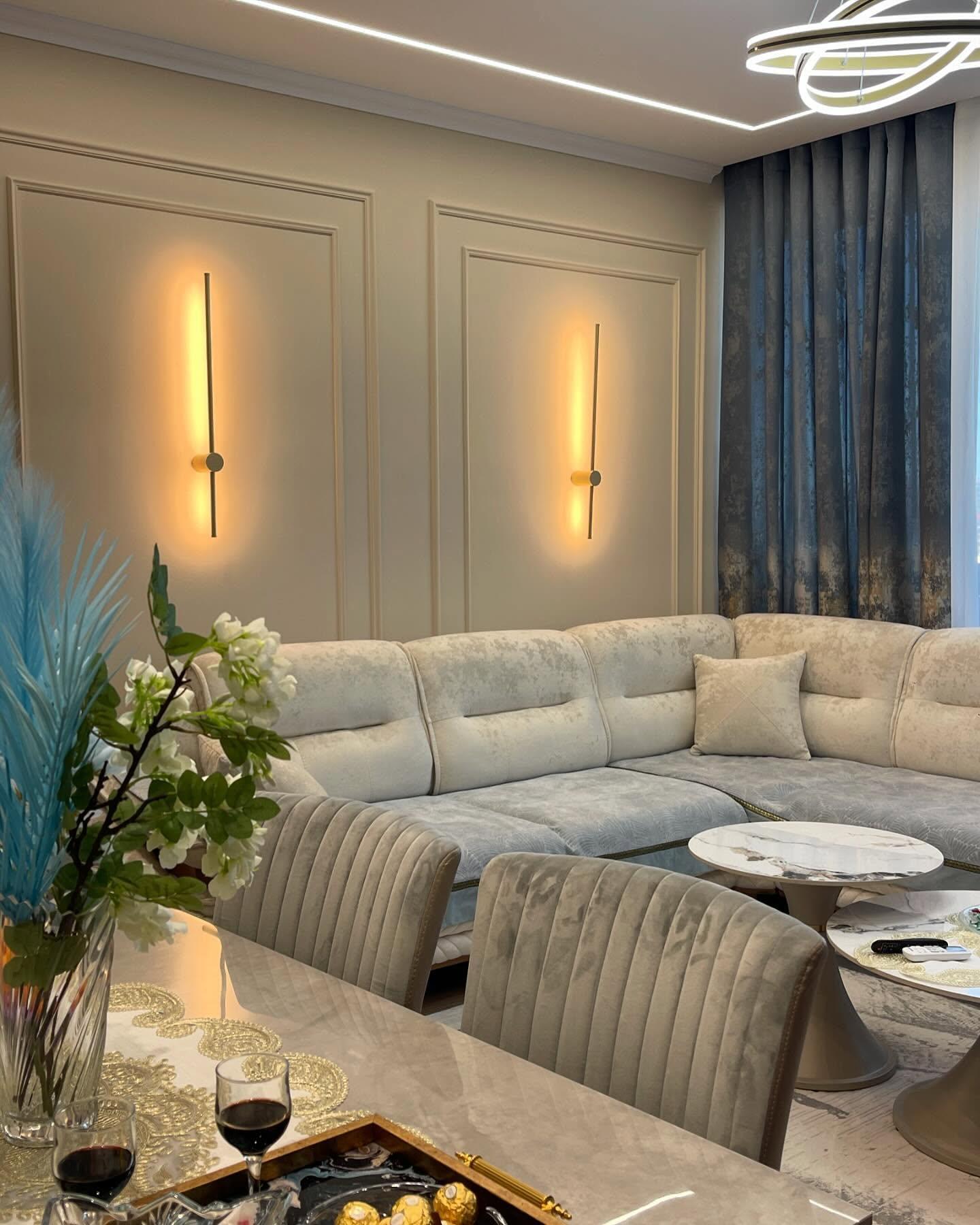 Living room with panelled walls, linear bronze wall lights, a large sectional sofa and a dining table in the foreground.